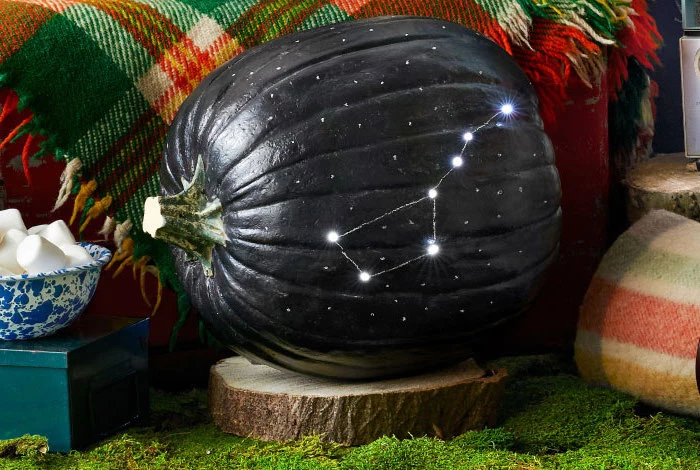 pumpkin carving ideas, pumpkin painted in black, constellation carved into it, sitting on a wooden log, on top of moss
