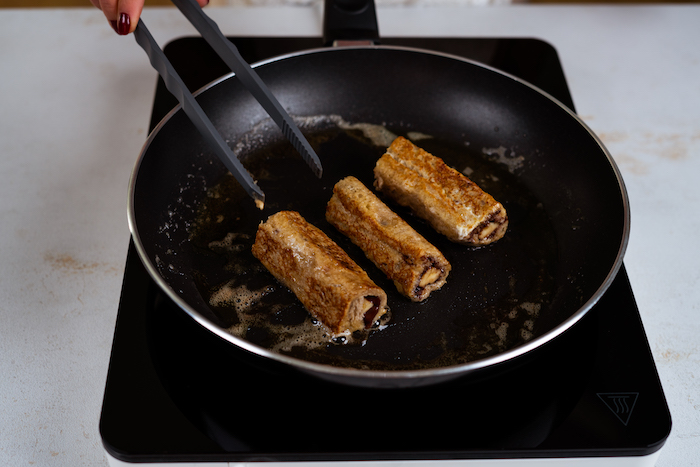 three bread roll ups, chocolate and bananas inside, french toast recipe, fried in butter in black sauce pan