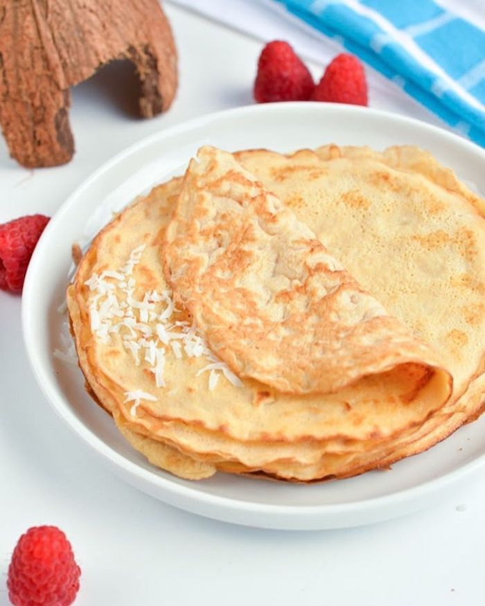 stack of crepes, keto breakfast, coconut flakes on top, white plate, raspberries scattered around