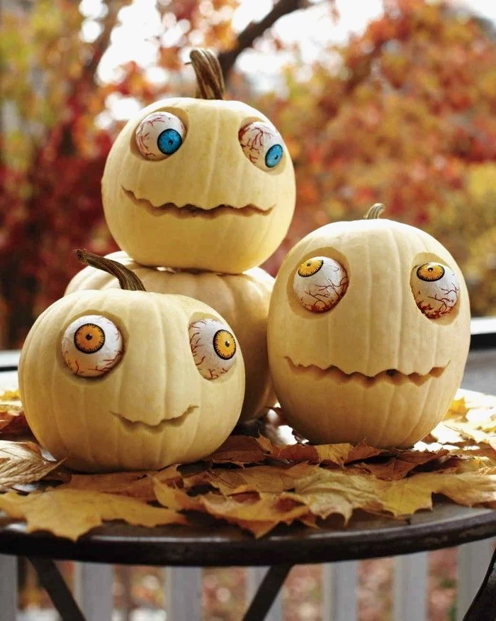 three white pumpkins, with large plastic eyes, pumpkin faces ideas, arranged on a black table, with fall leaves, pumpkin inside pumpkin carving patterns