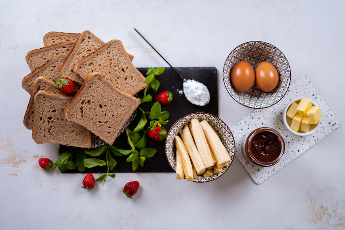 bread slices and strawberries, eggs bananas butter and chocolate in bowls, french toast, arranged on white countertop