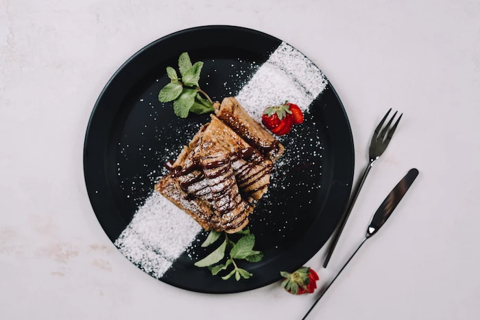 french toast, roll ups drizzled with chocolate, dusted with powdered sugar, strawberries on the side, placed on black plate