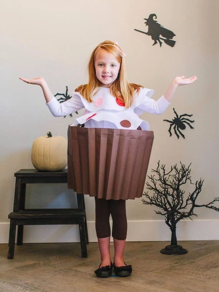 girl with blonde hair, dressed as a cupcake, made of paper, toddler girl costume, halloween decorations, in the background