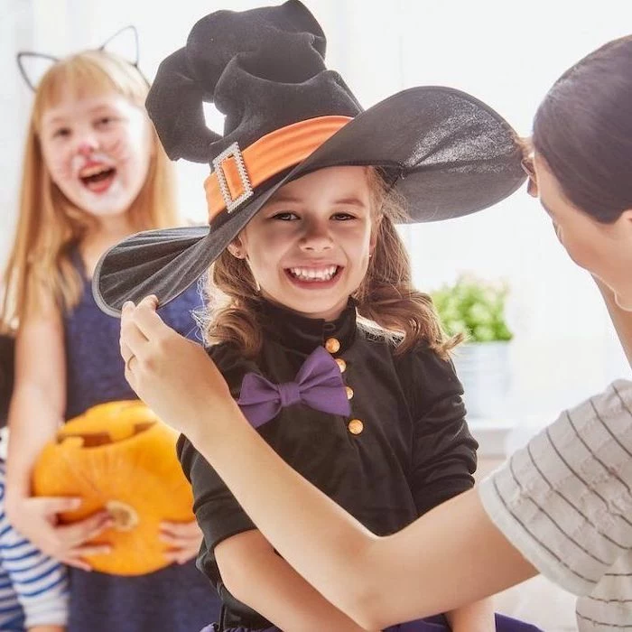 girl smiling, dressed as a witch, with large black hat, cute halloween costumes for girls, purple bowtie