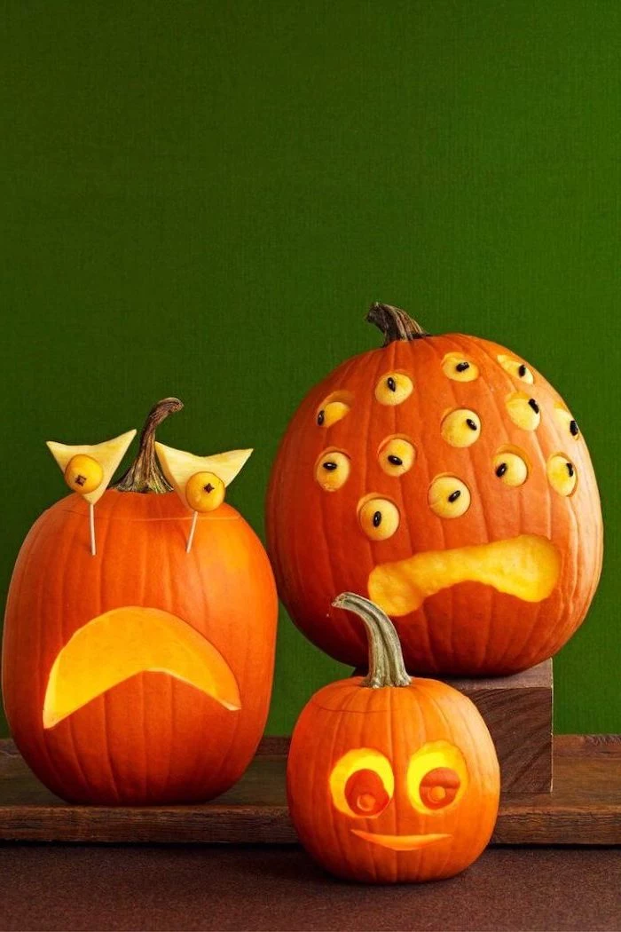 three pumpkins, with funny eyes, pumpkin faces ideas, arranged on a wooden crate, green wall