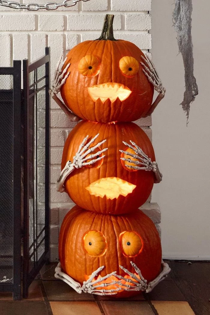 hear see say no evil, three pumpkins, stacked together, with skeleton hands, pumpkin carving, white brick wall