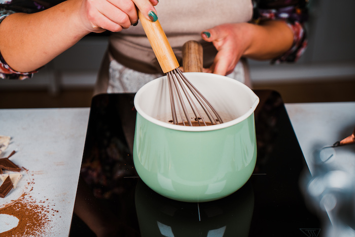 woman whisking chocolate mix in turquoise ceramic pot, homemade hot chocolate recipe, placed on white surface