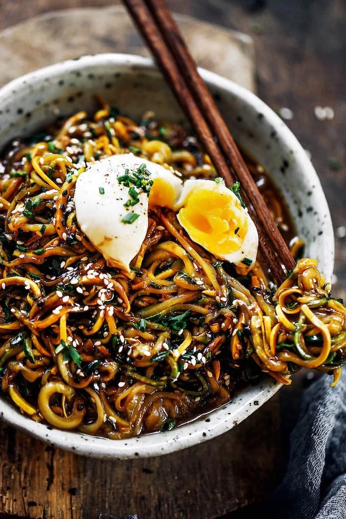 zucchini noodles, asian recipe, with sesame seeds, egg on top, in a white bowl, with wooden chopsticks