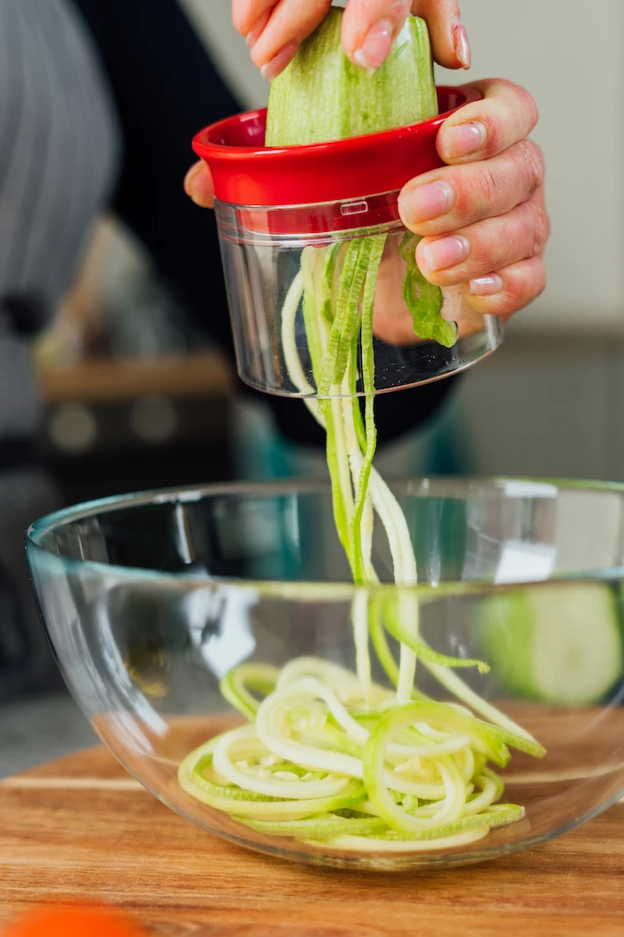 how to make zoodles zucchini being spiralised using spiraliser on top of glass bowl placed on wooden cutting board