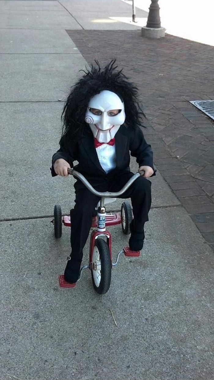 little boy, dressed as jigsaw, saw movie inspired, riding a tricycle, toddler boy halloween costumes, face mask