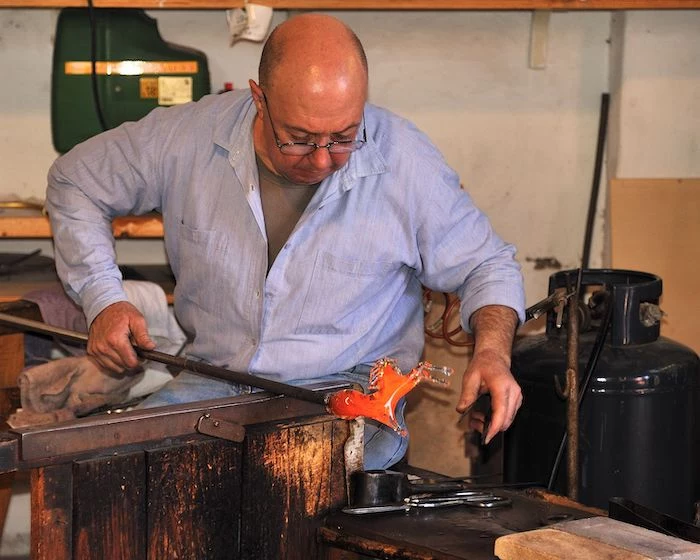 man making glass, wearing glasses, blue shirt, wooden table, different tools, murano glass chandelier