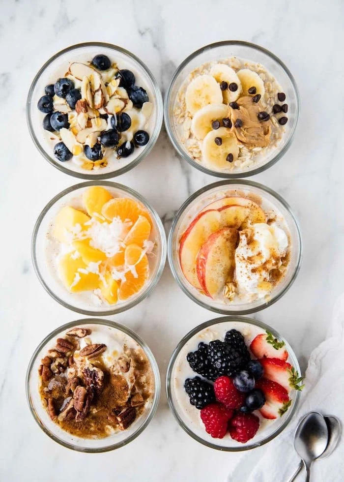 six glass bowls, filled with oatmeal, berries and bananas, apples and nuts, healthy lunch ideas for work, silver spoons