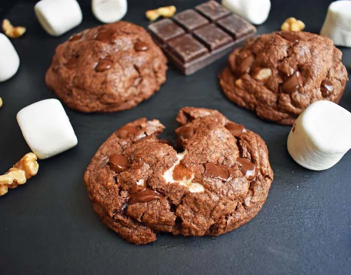 cocoa cookies, marshmallows inside, split in half, marshmallows scattered around, black table, chocolate chunk cookies