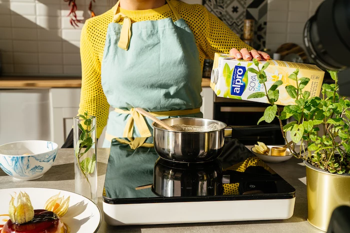 woman wearing yellow sweater and blue apron, pouring soya milk in pot over hot plate, creme caramel recipe