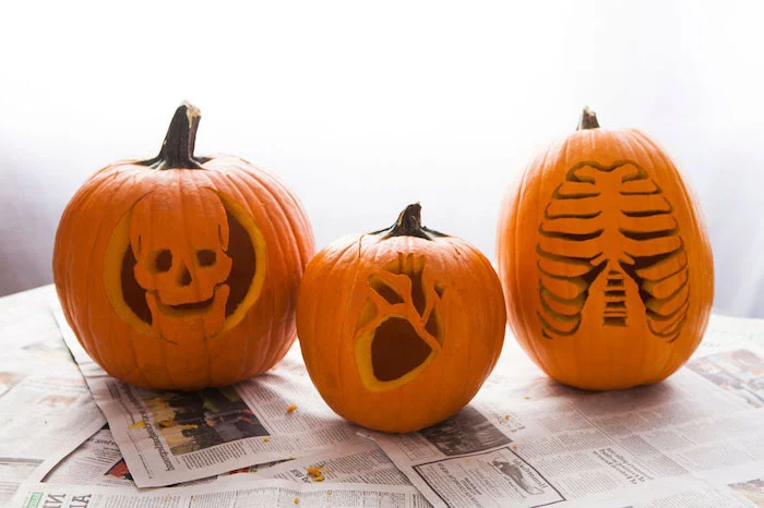 three pumpkins, skull and heart, rib cage, carved into them, pumpkin carving designs, arranged on top of newspapers