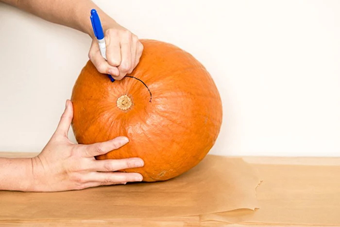 two hands, tracing a line, with a sharpie, onto a pumpkin, on a wooden table, jack o lantern faces, white wall