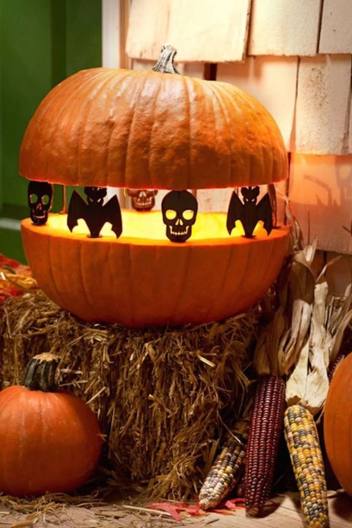 two pieces of a pumpkin, held together by figurines, skulls and bats, pumpkin carving patterns, on top of haystack
