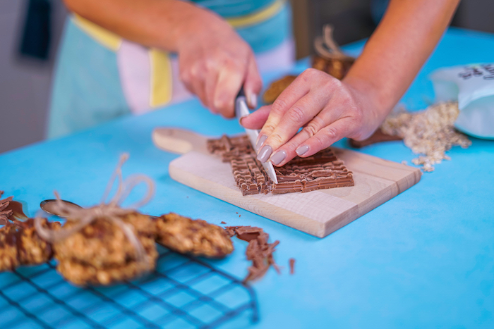 woman holding a knife, cutting chocolate, into pieces, chewy chocolate chip cookies, blue table, wooden cutting board