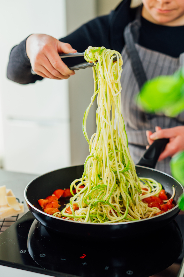 zoodles added to cooked cherry tomatoes in saucepan how to make zoodles being cooked with tongs