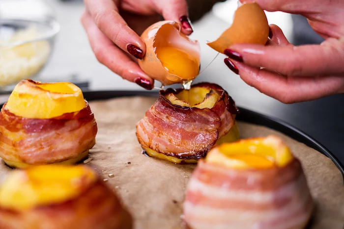 bacon slices wrapped around halved potatoes, baked potato volcanoes, filled with eggs, arranged on paper lined sheet pan