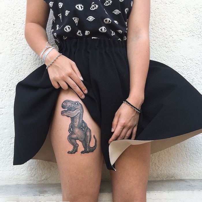 t rex dinosaur, thigh tattoos for women, woman wearing black skirt, black top, leaning on white wall