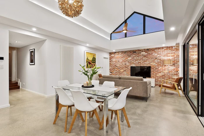 brick accent wall, glass table, white chairs, vaulted ceiling beams, grey sofa, white walls, tall sliding doors
