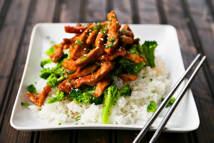 pork with sesame seeds, with broccoli and rice, in white plate, metal chopsticks on the side, what should i make for dinner, wooden table