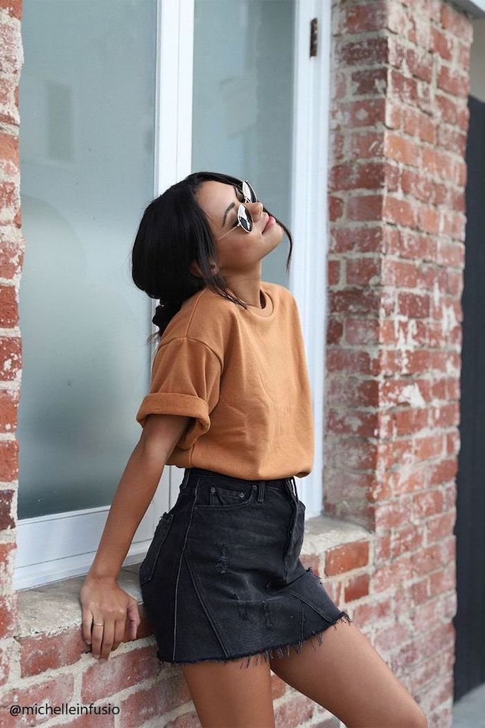 woman leaning on brick wall, wearing black denim skirt, orange t shirt and sunglasses, shoulder length hair, black hair in a bun