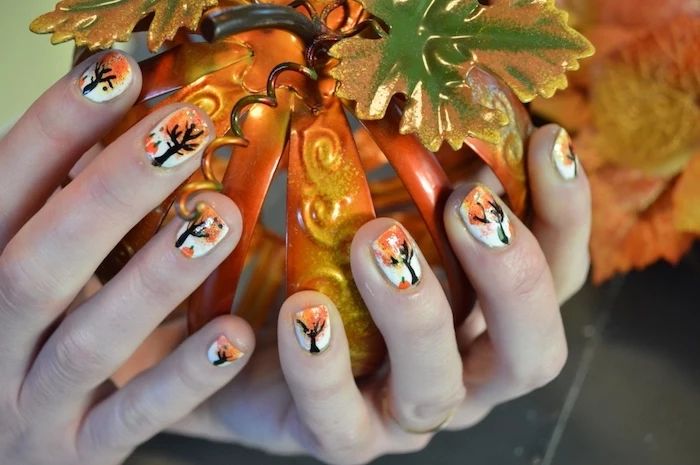 hands holding a ceramic pumpkin, white nail polish, fall nail ideas, trees with fall leaves, nail decorations