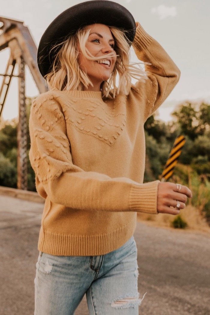 woman smiling, wearing a black hair, brown sweater and jeans, shoulder length bob, standing on a bridge