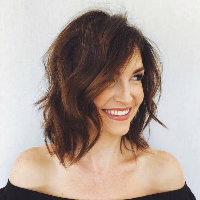 woman smiling, short to mid length hairstyles, wearing off the shoulder black blouse, brown wavy hair, white background