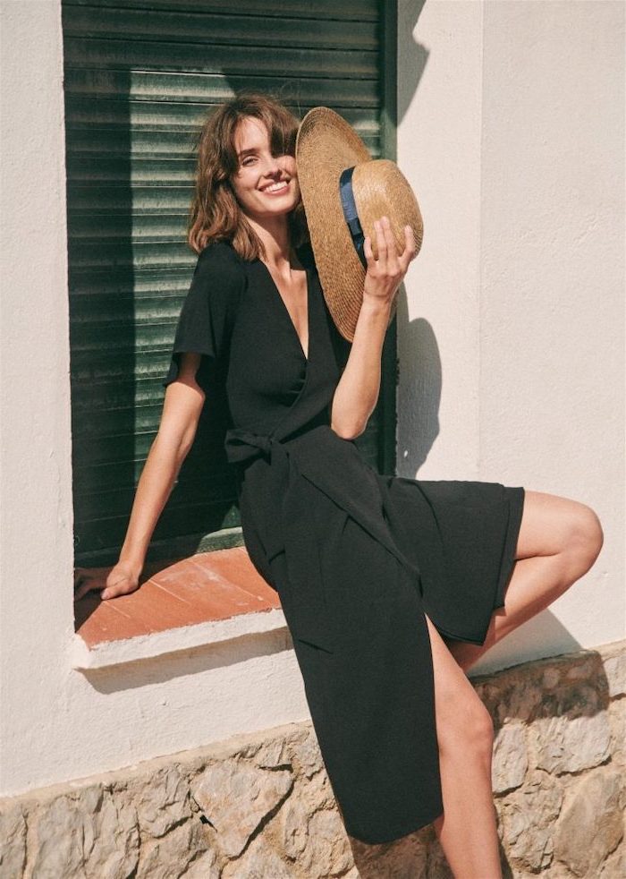 woman sitting on a window frame, wearing a black dress, holding a hat, short to mid length hairstyles, brown hair with bangs