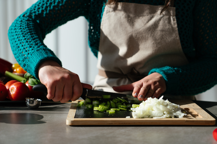 tomato soup recipe, celery and onion being chopped up with knife, on wooden cutting board, placed on grey countertop