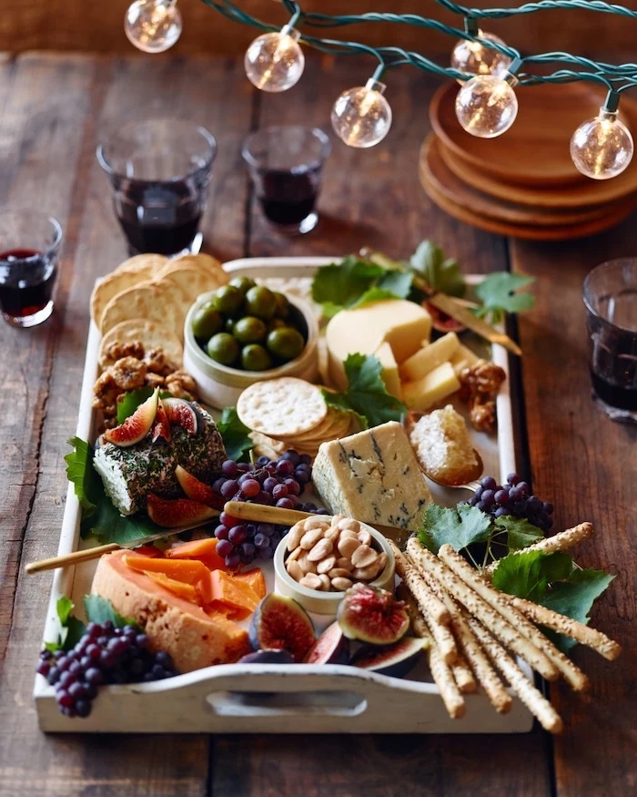cheese board with olives grapes, different types of crackers and pretzels, christmas appetizer ideas, wooden table