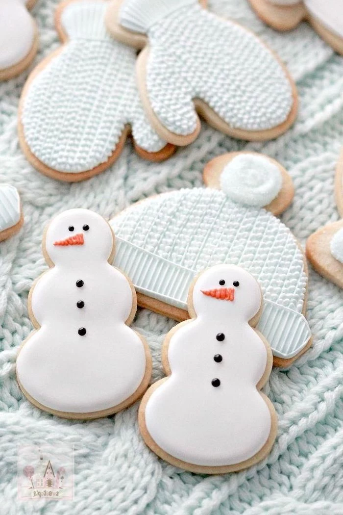 cookies in the shapes of snowmen mittens and beanie, cookie decorating icing, placed on white knitted cloth