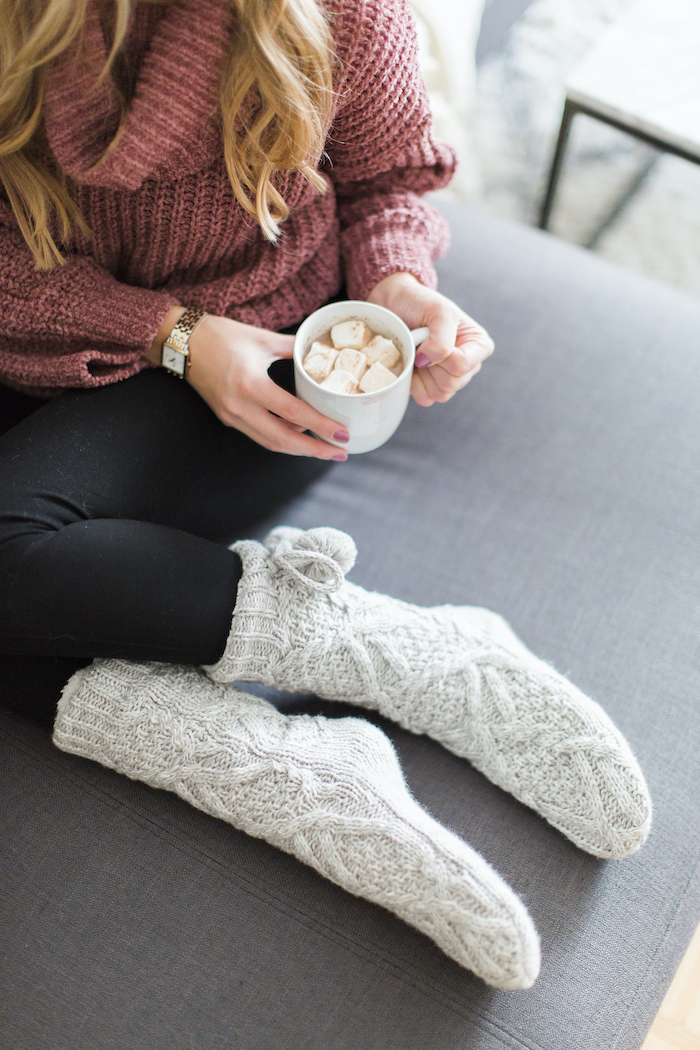 bonde woman wearing pink sweater and black tights, holding a cup with marshmallows, gifts for mom, grey knitted cozy socks