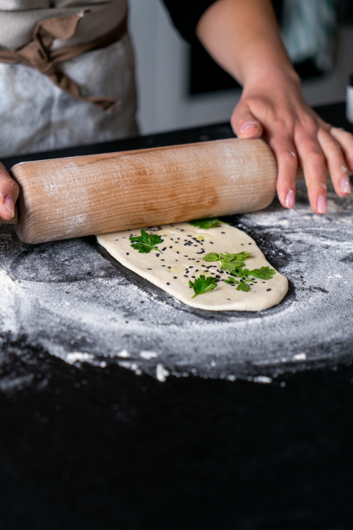 dough being rolled out, parsley and garlic, black sesame seeds placed on it, naan bread recipe, black surface covered with flour
