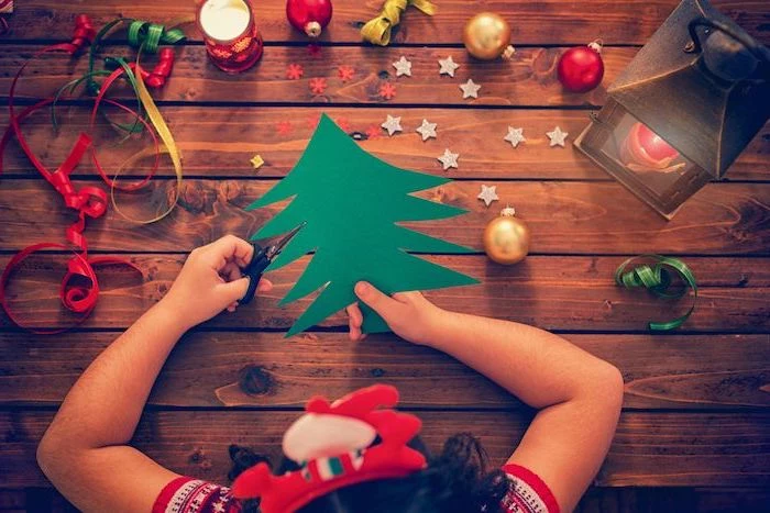 girl cutting a christmas tree out of green paper, christmas craft ideas for kids, baubles placed on wooden surface