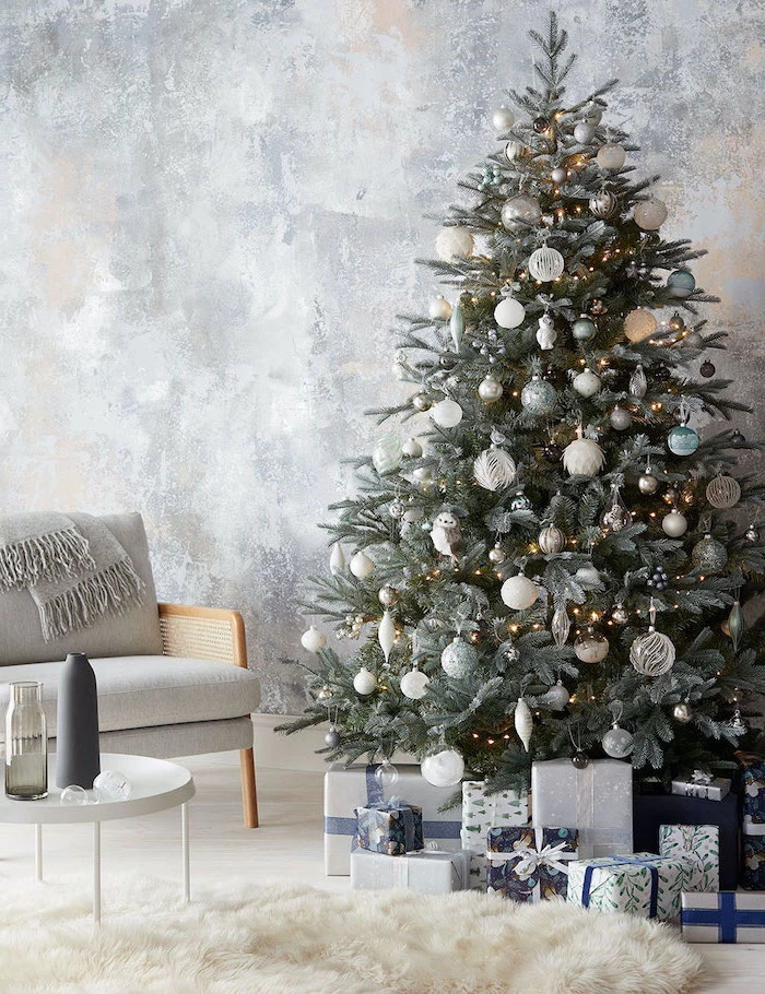 tree decorated with lights, white silver and gold ornaments, rose gold christmas tree, next to grey armchair, presents underneath