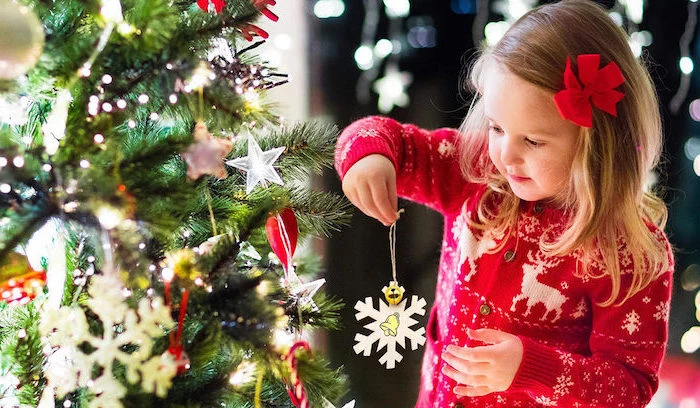 diy christmas ornaments for kids, little girl holding a handmade paper snowflake, next to a decorated christmas tree