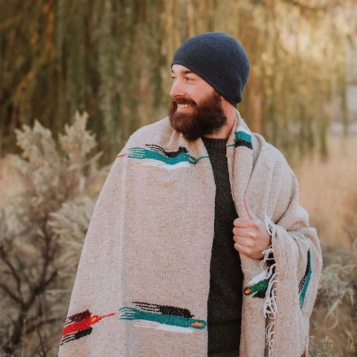 man with beard smiling, wrapped in a large cozy blanket, wearing black sweater and blue beanie, unique gifts for men