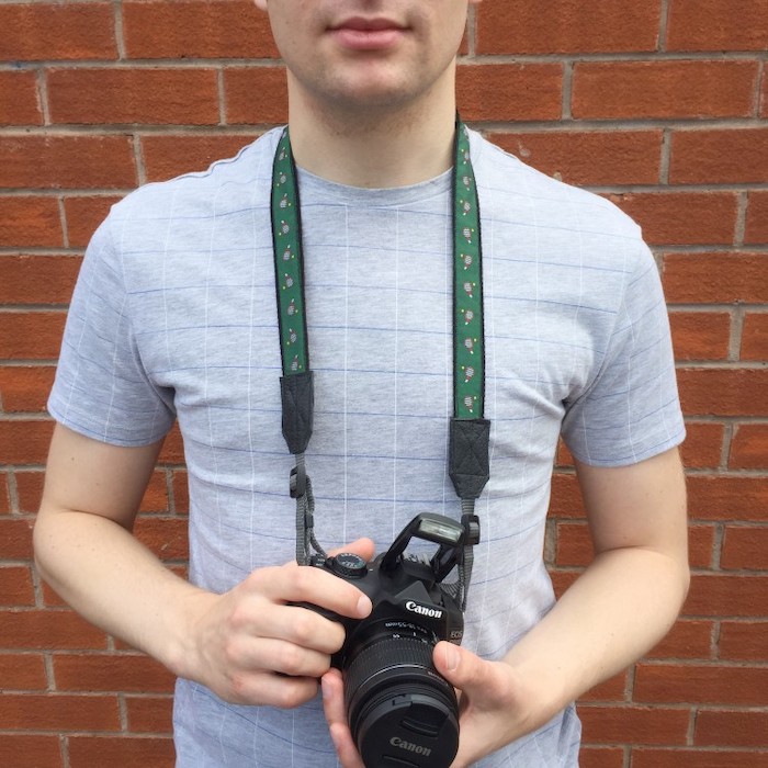 man holding a canon photo camera with green strap, step by step diy tutorial, wearing grey shirt, gift ideas for men, brick wall in the background