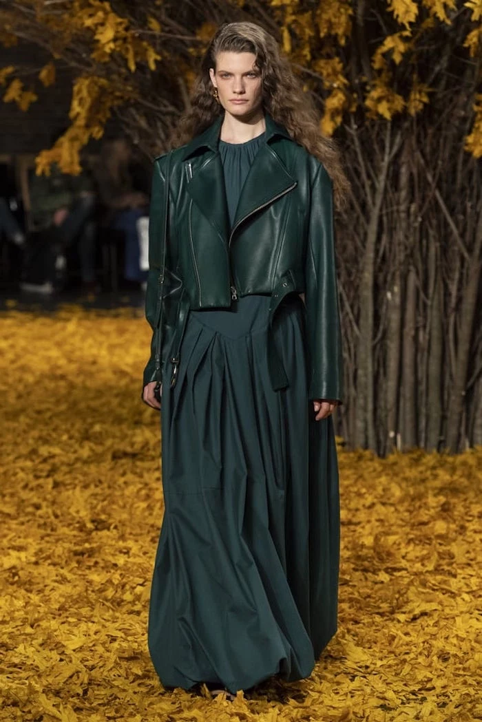model walking down a carpet of fall leaves, wearing dark green dress, fall fashion trends, dark green leather jacket