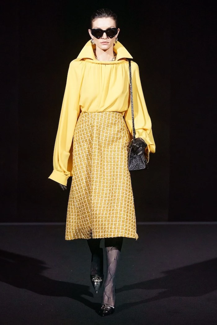 model walking down the runway, clothing trends, wearing long yellow skirt and blouse, black tights and shoes