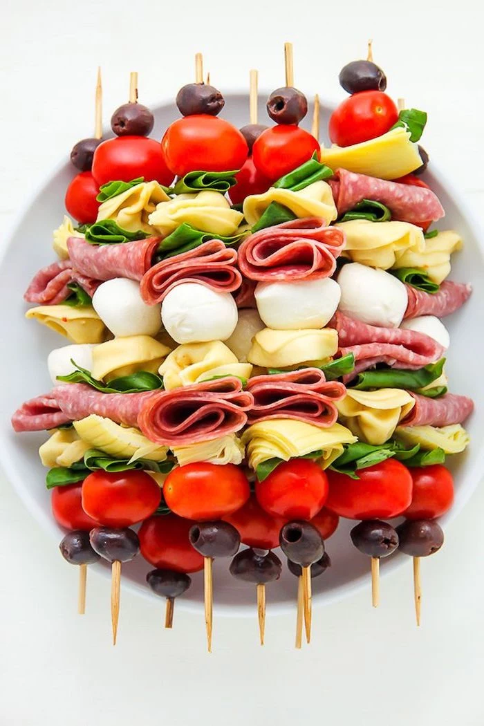 wooden skewers with meat, cheese and basil leaves, cherry tomatoes and olives, easy holiday appetizers, arranged on white plate