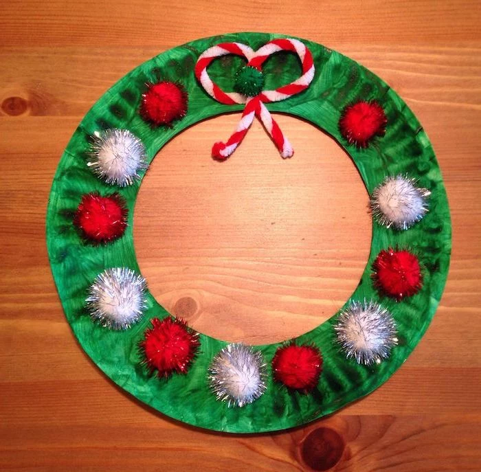 wreath made of paper plate, painted green with red and silver pompoms, diy christmas ornaments for kids, placed on wooden surface