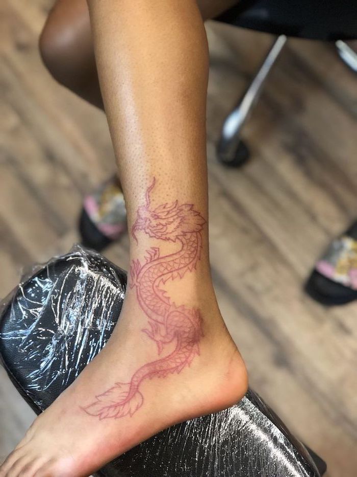 large red dragon, leg and ankle tattoo, what does a dragon symbolize, leg leaning on black leather chair, on wooden floor