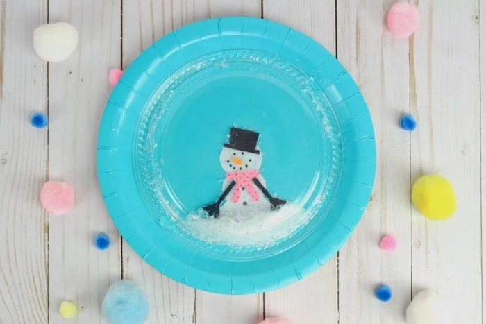 blue paper plate with snowmen drawn on it, plastic lid on top, faux snoq inside, diy ornaments for kids, placed on wooden surface
