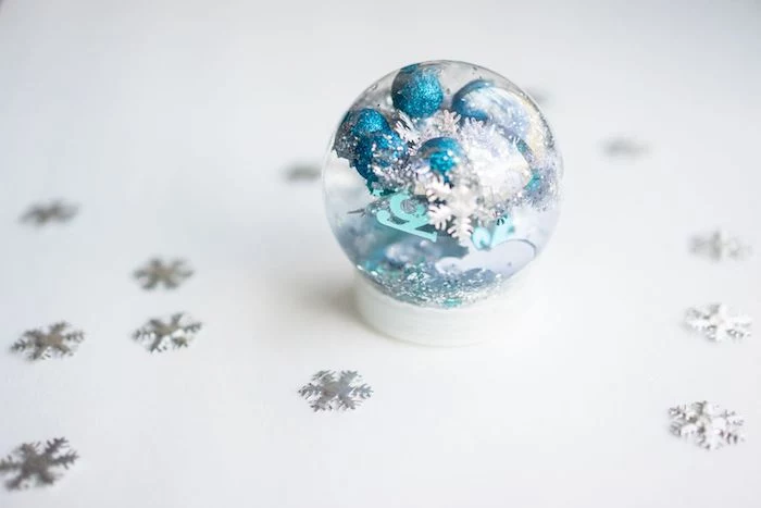 snow globe, with blue and silver pompoms, christmas ornament crafts, placed on white surface, silver snowflakes