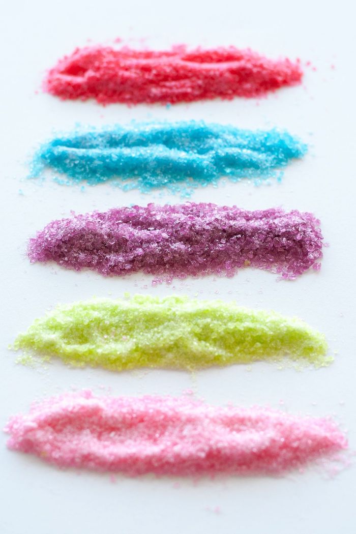 cookie icing recipe, sugar separated in 5 different piles, dyed in different colors, red blue purple green and pink, on white surface